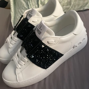 Valentino Garavani Rockstud sneakers💖 - Snygga vita Valentino Garavani Rockstud sneakers i skinn med svart band över foten dekorerat med svarta och silvriga nitar. Klassisk rund tå, vita snören och coola pyramidformade detaljer på sulan. Perfekta för dig som vill sticka ut med lyxig streetstyle.