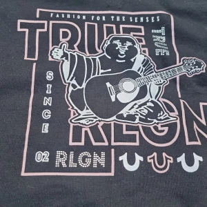 Svart croppad hoodie True Religion - Svart croppad hoodie från True Religion med ikoniskt Buddha-tryck och gitarr på bröstet. Tröjan har långa ärmar, huva och breda muddar. Snyggt tryck i vitt och rosa detaljer framtill. Har tvättnoppror på insidan men fortfarande skön att ha på sig :) Storlek S.