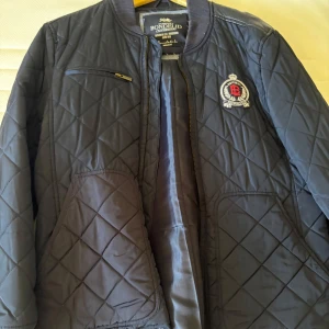 Quiltad blå jacka från Bondelid - Snygg quiltad blå jacka från Bondelid med emblem på bröstet och dragkedja framtill. Jackan har ribbad krage, lång ärm och praktiska fickor. Perfekt för dig som gillar klassisk stil med en sportig touch.