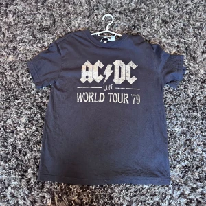 Svart AC/DC t-shirt från H&M - Svart t-shirt från H&M med AC/DC-tryck och texten 'Live World Tour '79' på bröstet. Klassisk rund hals och korta ärmar. Mjuk bomullskvalitet som är skön att bära. Perfekt för dig som gillar bandtröjor och vill ha en cool, avslappnad stil.