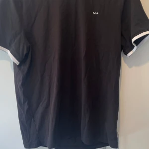 Svart t-shirt från Michael Kors - Svart t-shirt från Michael Kors med vit MK-logga broderad på bröstet. Klassisk passform med korta ärmar och vita detaljer vid ärmsluten. Tillverkad i mjuk bomull som känns skön mot huden. Perfekt för en clean och stilren look.