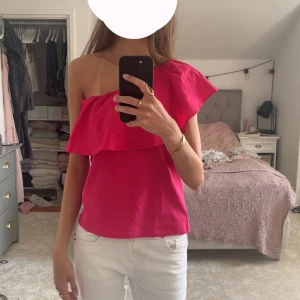 Rosa one shoulder volangtopp - Snygg rosa topp med one shoulder-design och bred volang längs överdelen. Toppen är ärmlös på ena sidan och har en smal axelrem på andra. Perfekt för dig som vill sticka ut med en färgstark och trendig look. Eftertraktad topp, som är i helt nytt skick med prislappen kvar!! 🩷(se bild) storlek 38-40 men passar även s-xs