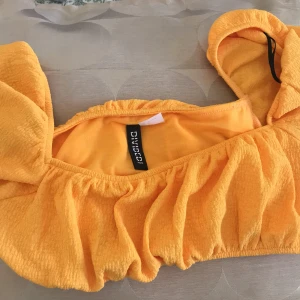 Gul one shoulder topp från H&M Divided - Superfin gul topp från H&M Divided med one shoulder-design och volangiga detaljer. Toppen är croppad och har en lätt strukturerad yta som ger en cool känsla. Perfekt för dig som vill sticka ut med färg och form i sommar.