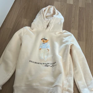 Beige Polo Bear hoodie från Ralph Lauren - Säljer en beige hoodie från Polo Ralph Lauren med ikoniska Polo Bear-trycket framtill. Tröjan har huva och är långärmad, perfekt för en chill och stilren look. Mjuk insida och klassisk passform. Storlek M.