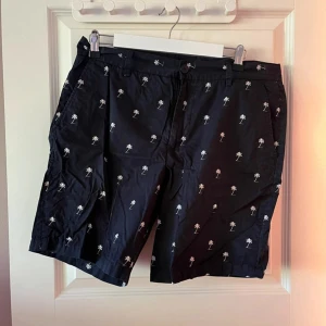 Blåa  shorts med palmtryck - Blåa shorts med vita små palmträd över hela tyget. Klassisk passform med fickor på sidorna och knappstängning. Perfekta för sommaren och chill dagar. Materialet känns lätt och svalt, troligen bomull. Snyggt och avslappnat mönster för en cool vibe.