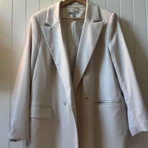 Beige kavaj med knappar - Stilren beige kavaj i storlek L med klassisk krage och dubbla knappar framtill! Kavajen har långa ärmar med tre dekorativa knappar vid ärmarna och två fickor🤗Materialet är slätt och ger en clean look, perfekt för att lyfta vilken outfit som helst💕