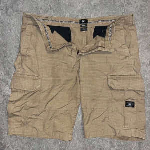 Beige cargoshorts från DC Shoes - Säljer ett par beige cargoshorts från DC Shoes i avslappnad passform. Shortsen har flera stora fickor med lock, dragkedja och bälteshällor. Materialet är bomull med diskret rutmönster i tyget. Perfekta för chill dagar och streetwear.