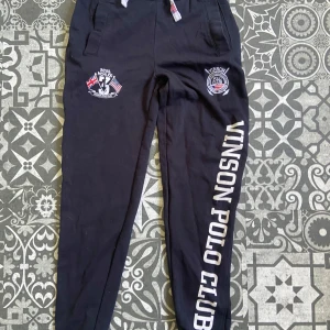 Svarta mjukisbyxor Vinson Polo Club - Svarta mjukisbyxor från Vinson Polo Club med broderade loggor och tryck längs benet. Byxorna har snörning i midjan med detaljer i rött och vitt, samt ribbad mudd upptill. Perfekta för en chill och sportig look.