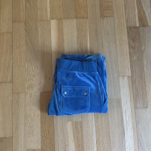 Blå velourbyxor Juicy Couture S - Blåa mjukisbyxor från Juicy Couture i mjuk velour med dragsko i midjan och raka ben. Byxorna har stora fickor fram, en bakficka med knappar och broderad JC-logga på ena fickan. Perfekta för en hemmakväll eller en snygg outfit! 💞
