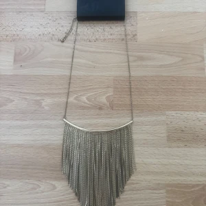 Ny. Guldigt franshalsband från H&M - Snyggt statementhalsband i guld från H&M med långa fransar av kedjor som hänger i en V-form. Perfekt för att lyfta vilken outfit som helst med en cool och trendig touch. Justerbar längd med kedja i nacken.