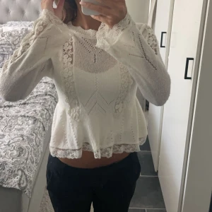 Vit spetsblus med volangdetaljer - Superfin vit blus med spetsdetaljer och volangkant. Blusen har långärmade ärmar med spets längs hela ärmen och blommiga broderier framtill. Den är lätt transparent och har en feminin, lite utsvängd passform. Perfekt för dig som gillar romantisk stil.