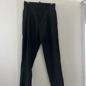 Svarta kostymbyxor - Klassiska svarta kostymbyxor med raka ben och snygga pressveck framtill. Byxorna har hög midja och stängs med dragkedja och knapp. Perfekta för en clean och stilren look.
