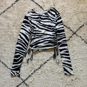 Zebramönstrad långärmad topp med snörning - Snygg långärmad topp med zebramönster i svart och vitt. Toppen har trendig snörning i sidorna som gör att du kan justera passformen och skapa en cool look. Perfekt för dig som vill sticka ut med ett djurmönster och gillar streetstyle.