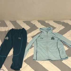 Marseille Tracksuit ljus och mörk blå - Säljer en ljus och mörkblå Marseille tracksuit från Puma med dragkedja och huva. Jackan har Vita och mörkblå detaljer och Marseille-logga på bröstet samt text på ryggen. Perfekt för fotbollsträning.