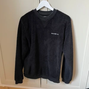 Svart fleece tröja från Woodbird - Mysig svart fleece tröja från Woodbird med rund hals och ribbade muddar. Perfekt för kyliga dagar. Tröjan har en diskret logga på bröstet.