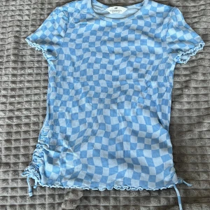 Blå och vit randig topp från H&M - Säljer en blå och vit randig topp från H&M med korta ärmar och ribbad struktur. Toppen har en unik snörningsdetalj på sidan och vågiga kanter vid ärmslut och nederkant. Perfekt för en avslappnad stil!