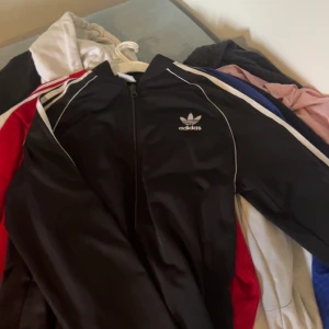 Svart träningsjacka från Adidas - Oanvänd tröja från adidas.                                   Snygg svart träningsjacka från Adidas med klassiska vita ränder längs ärmarna och dragkedja framtill. Perfekt för sportiga aktiviteter eller en avslappnad stil. Adidas-logga på bröstet ger en ikonisk touch.