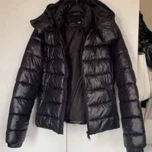 Svart dunjacka med huva - Säljer en svart dunjacka från lager 157 med huva och dragkedja framtill. Jackan har långa ärmar och två fickor med dragkedjor. Perfekt nu till våren och höst då den är lite tunnare. Knappt använd!