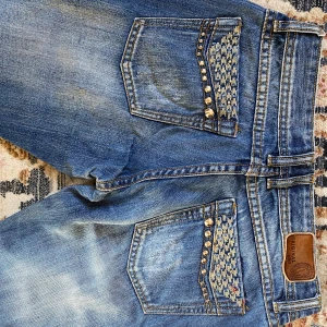 Lågmidjade jeans - Bredd ca 35 cm, längd ca 94, innerbenslängd ca 73. Snygga lågmidjade jeans med coola detaljer, köpta begagnat men passade ej, fint skick förutom fläckarr på baksidan av vänstra benet (synd bild 5) 