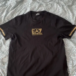 Svart t-shirt från Emporio Armani - Snygg svart t-shirt från Emporio Armani med guldfärgad logga på bröstet. T-shirten har korta ärmar med guldfärgade detaljer vid ärmsluten. Perfekt för en stilren look.