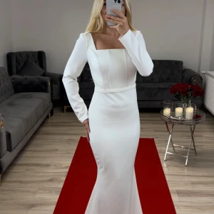 Vit långklänning med långa ärmar - Elegant vit långklänning med långa ärmar och en figurnära passform. Klänningen har en fyrkantig urringning och en vacker släp. Perfekt för formella tillfällen.