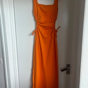 Orange ribbad klänning - Snygg orange ribbad klänning med knytning i midjan och en slits på sidan. Perfekt för en somrig look. Klänningen har en tight passform och är ärmlös med breda axelband.