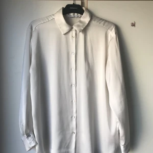Vit blus från Zara Basic - Elegant vit blus från Zara Basic med klassisk krage och knappar framtill. Blusen har långa ärmar och en stilren design som passar perfekt för både vardag och mer formella tillfällen.