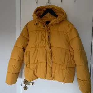 Gul dunjacka  - Gul dunjacka från laget 157. Jackan har dragkedja och knappar framtill samt snörning längst ner så att man kan dra åt. 