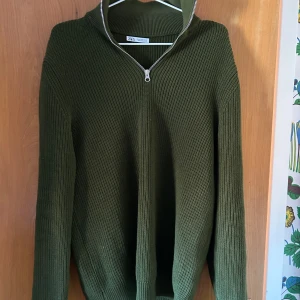 Olivgrön stickad tröja från Zara - Säljer en snygg olivgrön stickad tröja från Zara med dragkedja vid halsen. Tröjan har långa ärmar och en ribbad struktur som ger en stilren look. Perfekt för kyligare dagar!
