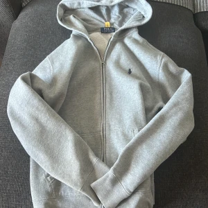 Ralph lauren zip up - Säljer en stilren grå hoodie från Ralph Lauren med dragkedja och en liten broderad logotyp på bröstet. Perfekt för en avslappnad look. Den har långa ärmar och en praktisk huva(det är storlek 170 men passar som XS) om ni har några frågor om plagget är det bara att skriva till mig -MVH Marko 