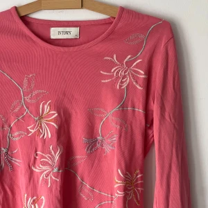 Rosa långärmad topp från Intown - Säljer en söt rosa långärmad topp från Intown med vackra broderade blommor i vitt och gult. Perfekt för att liva upp garderoben med lite färg och mönster. Toppen har en rund halsringning och är gjord i ett mjukt material.