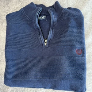 Mörkblå tröja med dragkedja från Chaps - Säljer en stilren mörkblå tröja från Chaps med dragkedja och broderad logga på bröstet. Tröjan har ett randigt mönster och är perfekt för kyligare dagar. Passar bra till både jeans och chinos. Passar även dem flesta som har storlek XL