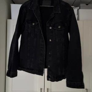 Svart jeansjacka från 157 - Säljer en svart jeansjacka från 157 i storlek M. Jackan har en klassisk design med knappar framtill och två bröstfickor. Perfekt för en casual look. Modellen är lite mer oversized och större