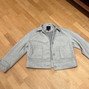 Grå kappa från Lager 157 - Säljer en stilren grå kappa från Lager 157 i storlek M. Jackan har en klassisk krage och knäppning framtill med stora fickor. Perfekt för höst och vår med sin eleganta look. Passar till många olika outfits!