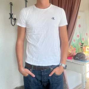 Vit t-shirt från Calvin Klein - Säljer en klassisk vit t-shirt från Calvin Klein med deras ikoniska logga på bröstet. T-shirten är liten i storleken och är mer en S/M. Tröjan har ett litet hål på magen som ej syns när man har den på.