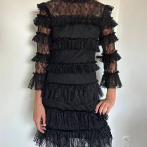 Svart spetsklänning med volanger - Elegant svart klänning i spets med volanger. Den har trekvartsärm och en tight passform som framhäver figuren. Perfekt för en festlig kväll.