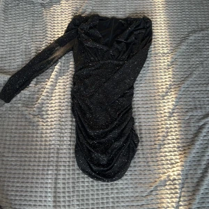 Svart glittrig klänning - Snygg svart klänning med glittrigt mönster och rynkade detaljer. Klänningen har en lång ärm och en åtsittande passform som ger en elegant look. Perfekt för en kväll ute eller en speciell tillställning.