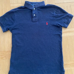 Marinblå pikétröja från Ralph Lauren - Säljer en stilren blå pikétröja från Ralph Lauren med röd broderad logga på bröstet. Tröjan har korta ärmar och en klassisk krage med två knappar. Perfekt för våren och sommaren.