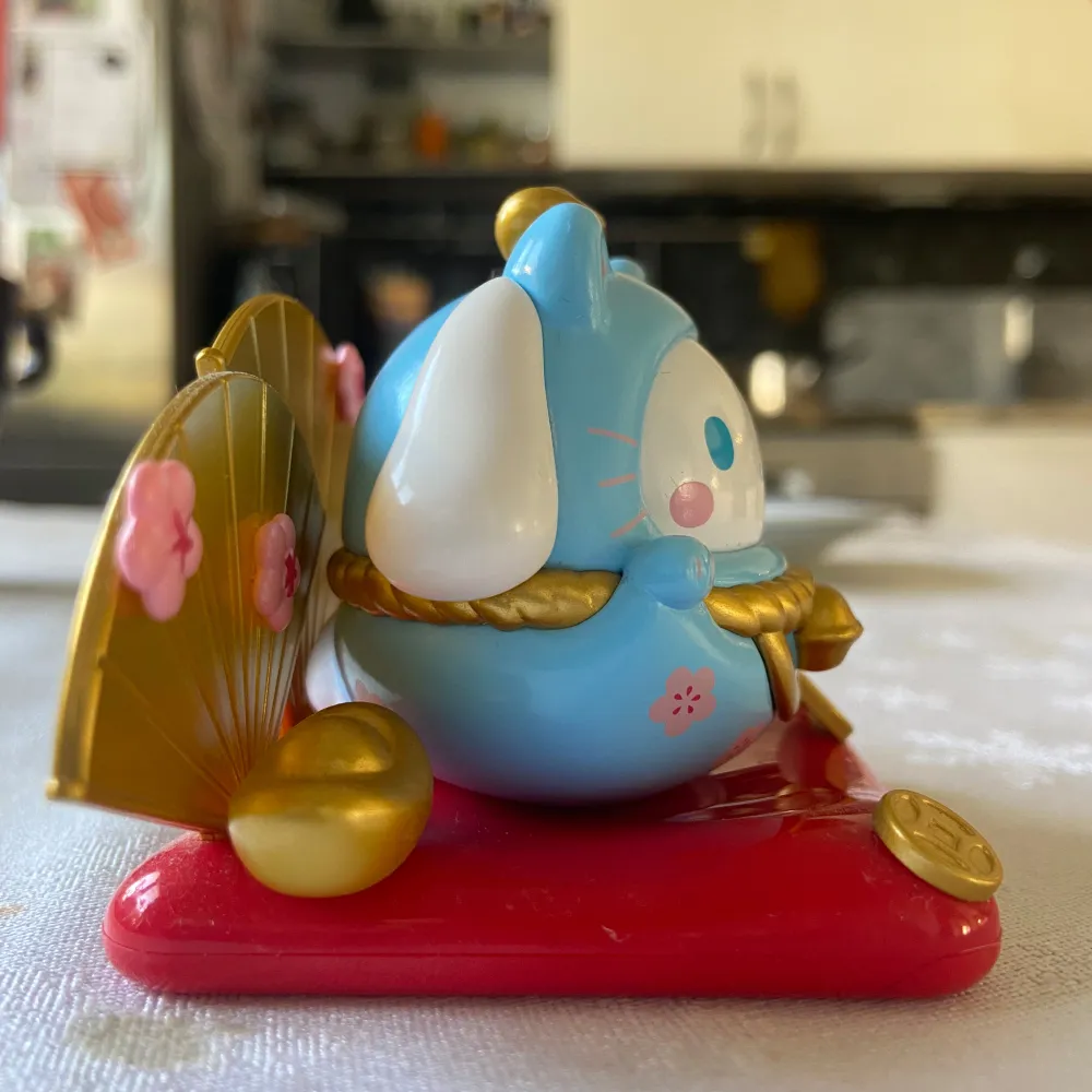 Charmig blå Cinnamoroll figur med kattöron och vita detaljer. Den har guldaccenter och rosa blommor, placerad på en röd bas med guldmynt. Perfekt som dekoration eller samlarobjekt.. Asusteet.