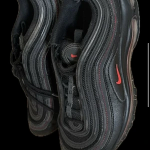 Svarta Nike Air Max 97 med röda detaljer - Snygga svarta Nike Air Max 97 med röda detaljer. Skorna har den ikoniska vågdesignen och en synlig Air-enhet i sulan för extra komfort. Perfekta för både stil och funktion. Skick 8/10