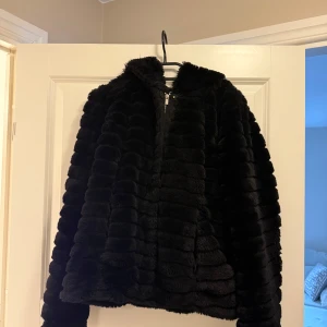 Svart pälsjacka med dragkedja och huva  - Säljer en snygg svart pälsjacka med dragkedja och luva. Hål i ena fickan men går att sy ihop snabbt. Passar både S och M