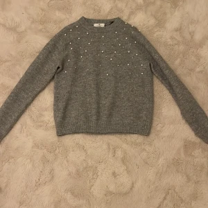 Grå stickad tröja med pärlor från H&M - Säljer en stilren grå stickad tröja från H&M dekorerad med pärlor på framsidan. Tröjan har långa ärmar och ribbade kanter vid ärmslut och nederkant. Perfekt för kyligare dagar! 🌟