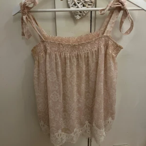 Beige blommig linnetopp med spetsdetaljer - Söt beige linnetopp med blommigt mönster och spetsdetaljer längst ner. Toppen har knytbara axelband och en luftig passform.
