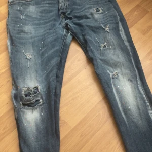 Blå jeans med slitningar - Säljer ett par blå jeans med coola slitningar och en avslappnad look. Perfekta för en casual stil med en touch av edge. De har en klassisk femficksdesign och knappgylf.