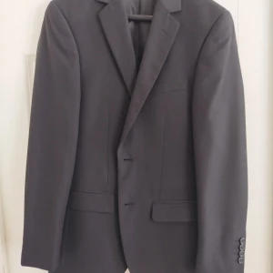 Svart kavaj från Dressmann - Snygg svart kavaj från Dressmann i slim fit 44. Använd en gång. Perfekt för en stilren look. Kavajen har klassiska slag och knappar framtill.
