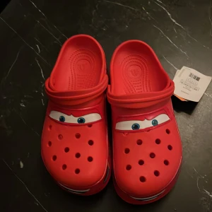 Röda Crocs med Blixten McQueen-motiv - Säljer ett par röda Crocs med motiv av Blixten McQueen från filmen Bilar. Skorna har en klassisk Crocs-design med hål för ventilation och en justerbar hälrem. Perfekta för alla små fans av Blixten McQueen! Storlek 45-46, helt oanvända. Säljer då de är för stora.