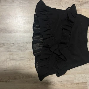 Svart kortkjol med volang - Snygg svart kortkjol med volangdetaljer. Perfekt för en stilren look. Kjolen har en omlott-design som ger en extra touch av elegans.