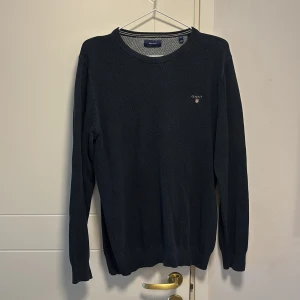 Mörkblå tröja från GANT - Säljer en stilren mörkblå tröja från GANT med rund halsringning. Tröjan har långa ärmar och ett diskret märke på bröstet. Perfekt för en klassisk och avslappnad look.