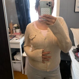 Beige/rosa kofta med knappar - Säljer en mysig beige kofta med knappar framtill och ribbade detaljer. Koftan har långa ärmar och en rund halsringning. Perfekt för kyliga dagar! Merket ar Guess