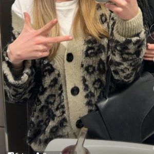 Beige och svart leopardmönstrad kofta - Säljer en mysig beige kofta med svart leopardmönster. Koftan har stora svarta knappar och ribbade muddar vid ärmsluten. Perfekt för att hålla sig varm och stilren.